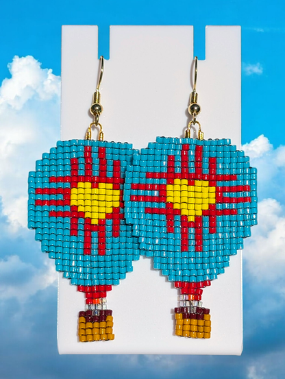 A pair of beaded Zia heart hot air balloon earrings over a cloudy, blue sky background. The earrings are mainly turquoise with the Zia shape in red and a yellow heart center.