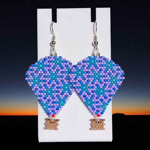 A pair of beaded lavender, blue, and turquoise hot air balloon earrings over a break of dawn background.