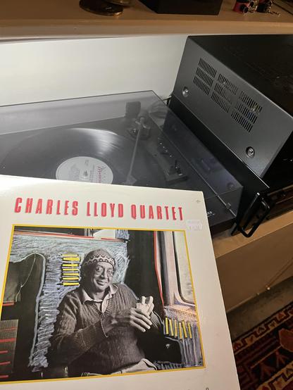 This photo shows another Charles Lloyd Quartet vinyl album, this time displayed on a turntable setup with a different aesthetic than the first image.

The Album:Charles Lloyd Quartet Montreux '82 record with a white cover featuring bold red text at the top. The cover shows a black and white photograph of Charles Lloyd himself, wearing glasses and a knit cap, smiling warmly at the camera. The image is framed with colorful geometric accents - yellow borders and striped patterns in the corners that give it a distinctive graphic design style.

The Turntable: A Marantz record player with a clear acrylic dust cover sits on a shelf. An adjacent tuner has a sleek black body with ventilation grilles visible on the side panel. A vinyl record is mounted on the platter, and you can see the tonearm and cartridge in playing position.

Setting: The turntable is positioned on a light-colored wooden cabinet against a white wall. There are some items visible in the background on the upper shelf, including other audio equipment.

Overall vibe: This is a clean, minimalist setup that contrasts with the more eclectic scene from the first photo, showing someone's dedication to playing and enjoying vintage jazz recordings on modern equipment.