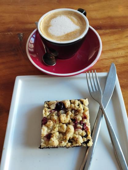 Una taza de café oscuro con arte latte en forma de corazón en la espuma, posada sobre un plato rojo brillante. A su lado, en primer plano, hay un trozo cuadrado de postre con una base oscura y una capa superior de crumble dorado con trozos de frutos rojos (arándanos y frambuesas). El postre está en un plato rectangular blanco, y a su derecha hay un tenedor y un cuchillo de metal plateado listos para ser usados. Todo se encuentra sobre una mesa de madera clara con vetas visibles.