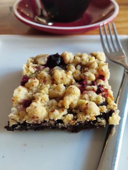 Un primer plano de una pieza de postre cuadrada sobre un plato blanco. La base es oscura, similar a un brownie de chocolate, y está cubierta con una capa gruesa de crumble dorado. En el crumble se aprecian arándanos enteros de color azul oscuro y trozos de frutos rojos, posiblemente frambuesas. A la derecha del postre, hay un tenedor plateado. En el fondo desenfocado, se distingue una taza oscura sobre un platillo rojo oscuro, con una cuchara dentro.