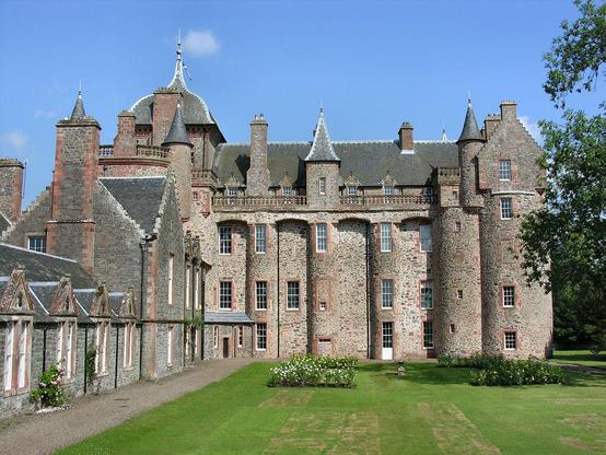 Thirlestane Castle. The image shows a view across a garden to a highly decorative wing of a castle built of grey stone with pink stone details and facings. There are turrets and chimneys along the roof, plus a walkway with a balustrade. There is another range on the left of the image, rather lower and plainer in style. The sky is mainly blue.