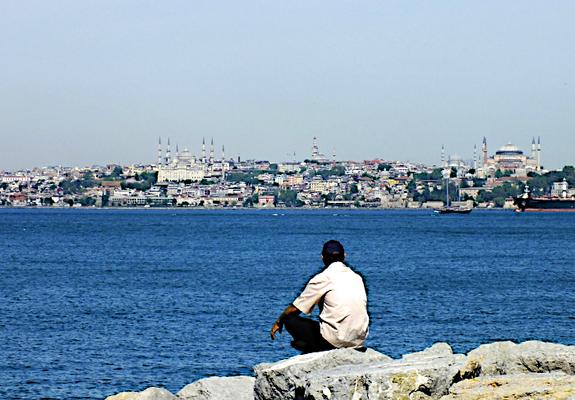 Foto van gehurkte man die over het blauwe water van de Bosporus naar het Europese deel van Istanbul kijkt