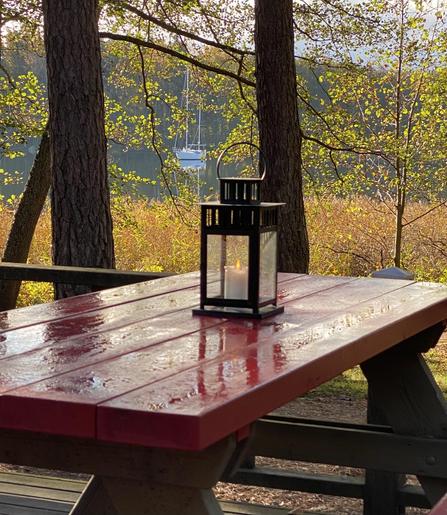 Puisista lankuista tehdyllä ulkopöydällä on kynttilälyhty ja sulaneesta kuurasta jäänyttä kosteutta. Taustalla männynrunkoja, tervaleppiä ja kellastunutta ruovikkoa sekä keskelle pientä lahtea ankkuroitu kaksimastoinen purjevene.