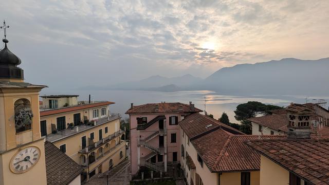 This elevated shot shows a church steeple on the left and several buildings with terracotta roofs in the middle ground. In the background, a large lake surrounded by a mountain range stretches out. The sun breaks through the clouds in the sky, reflecting its light off the water's surface.
Diese Aufnahme aus erhöhter Position zeigt links einen Kirchturm und im Mittelgrund mehrere Gebäude mit Terrakotta-Dächern. Im Hintergrund erstreckt sich der Lago Maggiore, der von einer Bergkette umgeben ist. Die Sonne bricht durch die Wolken am Himmel und ihr Licht spiegelt sich auf der Wasseroberfläche.