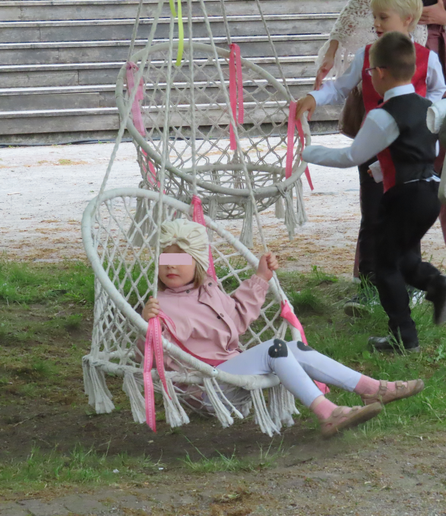 A Tallinn, lors d'une fête, une petit fille habillée en blanc et rose se balance dans une balançoire-siège suspendue blanche et décorée de rubans roses, derrière, deux petits garçons faisant partie d'une chorale en tenue de concert (ou traditionnelle ?) jouent avec une autre balançoire.
In Tallinn, during a festival, a little girl dressed in white and pink swings on a white swing seat decorated with pink ribbons. Behind her, two little boys who are part of a choir in concert (or traditional?)  play with another swing.