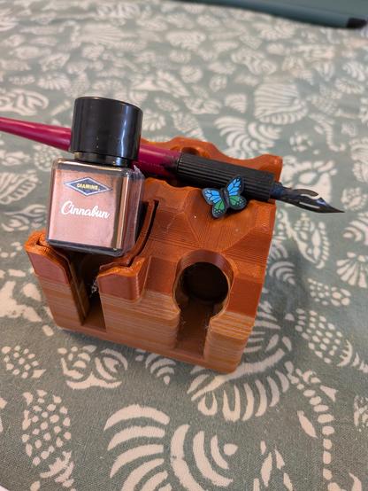 Against the back drop of a shell themed tablecloth a Cinnabun ink bottle sits upon an inkstand, with a dip pen, and a tiny Lego butterfly in blue.