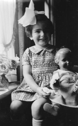 Vintage black and white photo of a smiling young girl wearing a large bow on their head and seated next to a window, holding a doll.