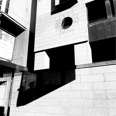 A black and white photograph of a textured building facade featuring a circular window, geometric shadows, and a bus stop sign.