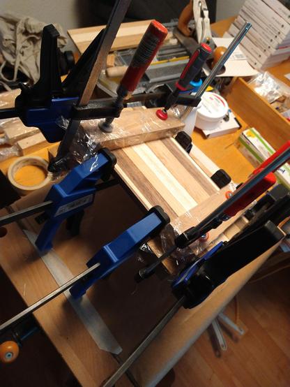Blick auf eine verspannte Leimholzplatte auf einer mobilen Werkbank. Verschiedenfarbige Holzstreifen werden mit vier Spann- und vier Schraubzwingen nebst Holzzulagen zusammengespannt, bis der Leim ausgehärtet ist. Um die Konstruktion herum sehen wir verschiedene andere Holzwerkzeuge.