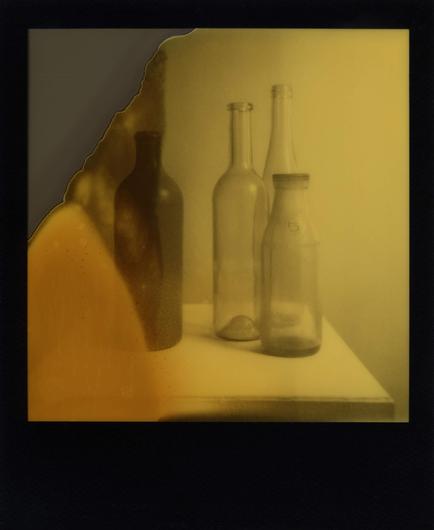 Ein Polaroidfoto mit schwarzem Bildrand zeigt drei Glasflaschen rechts und eine Tonflasche links in gelb-grauen Farbtönen. Auf der linken Bildseite ist ein Bildanteil nicht entwickelt und es hat sich eine orange bis graue Verfärbung mit kleinen Flecken gebildet.