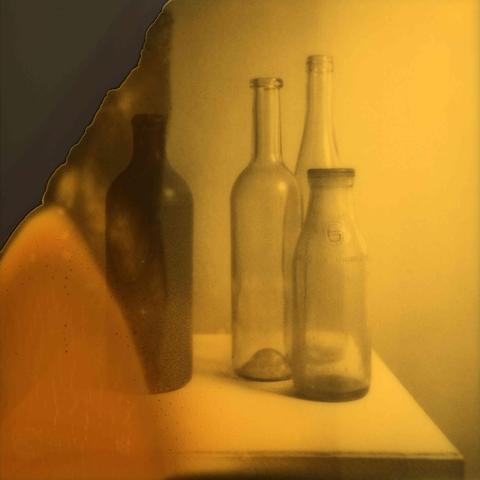 Ein Polaroidfoto zeigt drei Glasflaschen rechts und eine Tonflasche links in gelb-grauen Farbtönen. Auf der linken Bildseite ist ein Bildanteil nicht entwickelt und es hat sich eine orange bis graue Verfärbung mit kleinen Flecken gebildet. Dies ist der quadratische Bildausschnitt ohne Bildrand.