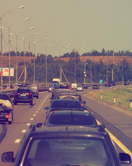Photo of a two-line (in one direction) interstate road with some cars on it. The road is turning right on the background.
Next to the road there is a hill with an yellow grass and the green trees.
Between the lanes and on the opposite lane there are light poles. On the right there are black and white boundary posts and the green grass.
Cars on the road mostly has black or grey color.
