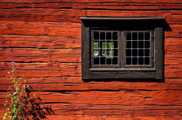 Red wooden wall with visible horizontal planks and a centrally positioned, dark-framed window featuring a grid pattern of small square panes. Reflection of greenery in the window panes. A flowering plant with pink blossoms grows at the bottom left corner.