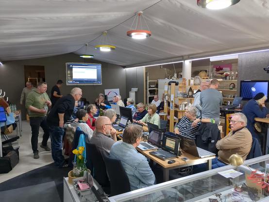 Ein großer Raum mit einem großen Fernseher an der Stirnseite. Im Raum stehen einige Tische, an denen Menschen mit ihren Notebooks und PCs sitzen.