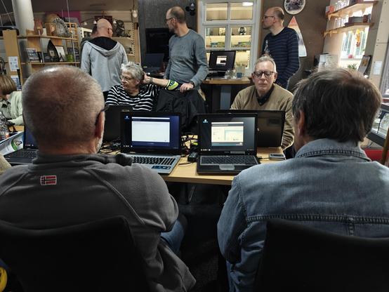 Mehrere Menschen sitzen vor ihren Notebooks. Im Hintergrund stehen weiterer Personen.