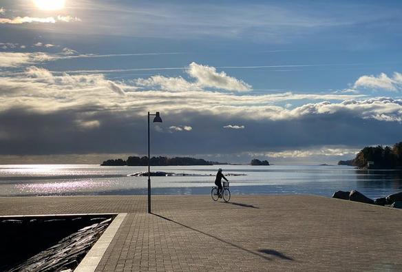Pyöräilijä lähtee liikkeelle tasaiselta laattapäällysteiseltä aallonmurtajalta. Taustana meri, metsäisiä saaria ja kaukana avomeren yllä pilviä. Aurinko kuvan vasemmassa yläkulmassa, vastavalo heittää pyöräilijän ja katulyhdyn varjot laatoitukselle.