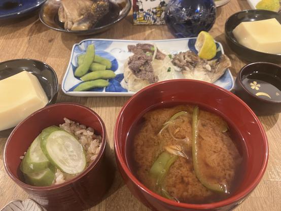 A Japanese meal displayed on a wooden table, featuring a red bowl of miso soup with green onions, a bowl of rice topped with cucumber slices, and various dishes including edamame, a plate of sliced beef and vegetables, and an egg tofu.