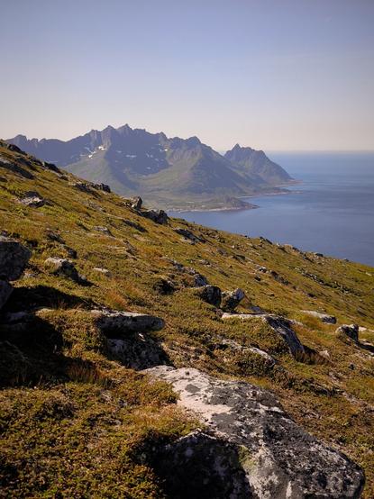 The image depicts a rugged, mountainous landscape with a clear blue sky. In the foreground, there is a grassy hillside with scattered rocks, covered in patches of green moss and grass. The hill slopes downward towards a body of water, likely a fjord, with a coastline visible in the distance. The mountains in the background are steep and jagged, with some snow patches visible on their peaks, indicating a high altitude. The water appears calm and reflects the blue of the sky, with a small settlement or village visible along the coast. The overall scene is serene and showcases the natural beauty of a mountainous region near the sea.