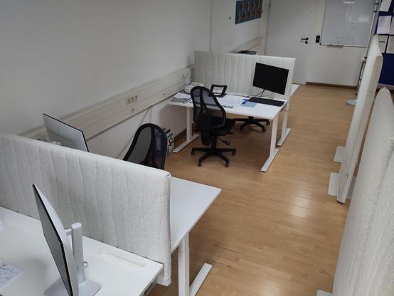 Büro-ähnlicher Raum mit weißen Schreibtischen und Bildschirmen , die mit Schall-dämpfenden grauen Elementen akustisch von einander isoliert sind.