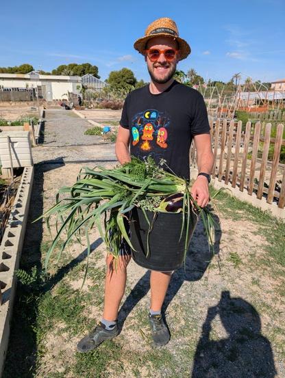 Hombre sonriente vestido con camiseta negra con dibujos de los fantasmas de pactan sentados alrededor de una hoguera, pantalones cortos, gafas de sol naranjas y sombrero de paja, sujeta un cubo de plástico negro repleto de hortalizas, rodeado de bancales y un cielo azul y despejado.
