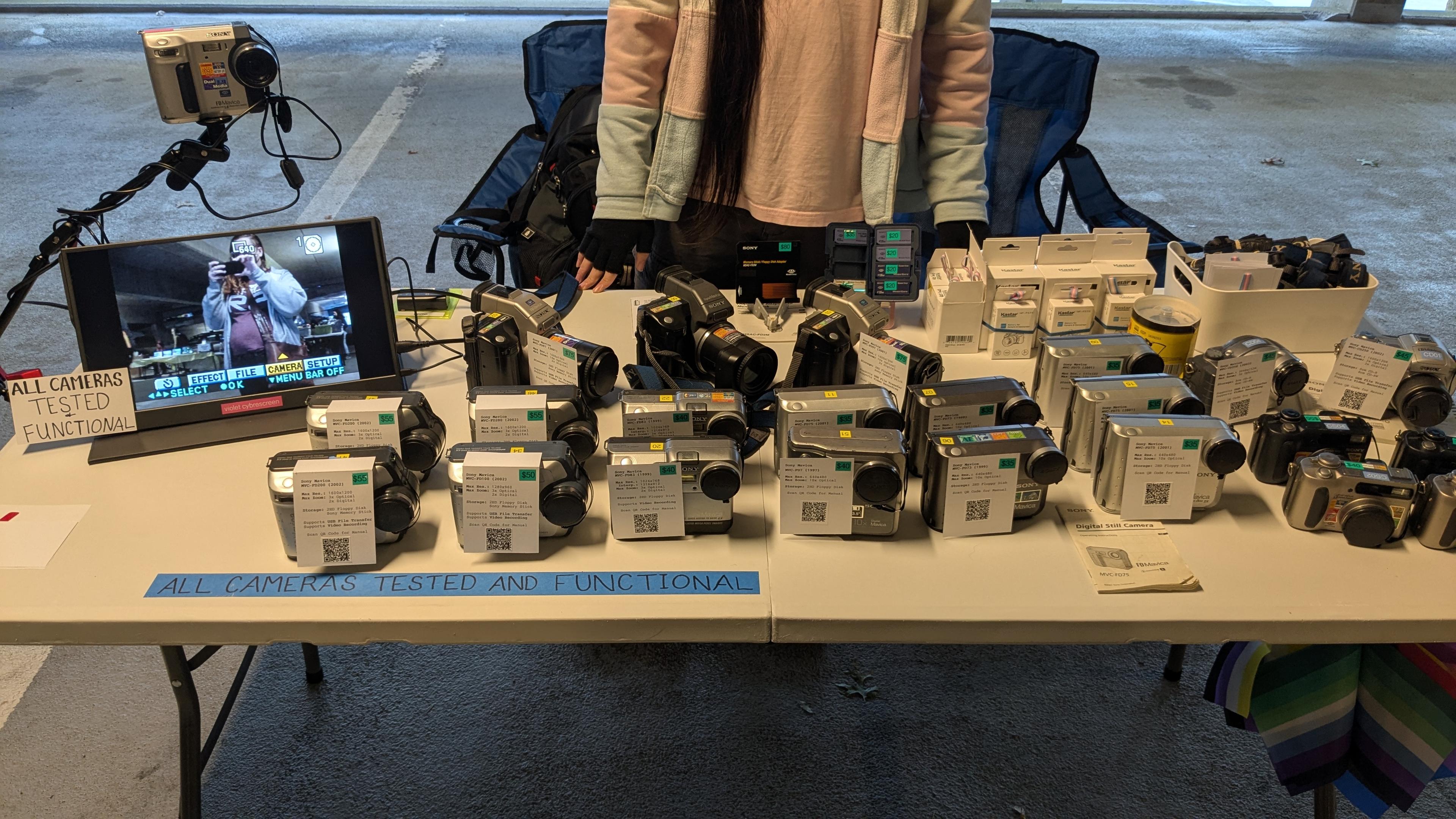 18 Sony Mavicas of various types including disc and floppy, and 4 CyberShots are laid out on a table, with signs saying "all cameras tested and functional". There's a 19th Mavica connected to a tablet showing me taking the picture.