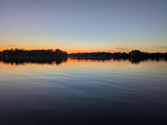 Sunset over the water with a clear sky. The sky and trees from the distance shoreline are mirrored in the calm water.