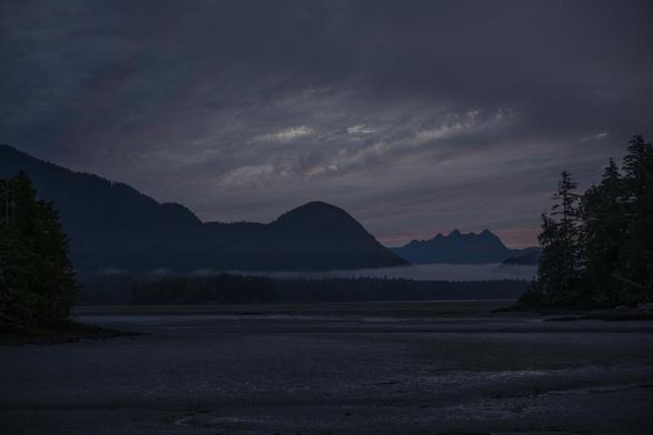 Dawn coming up over mudlfats (foreground), islands and deeper water (mid distance), and mountains (further back). 

Light just starting to come into the sky.

Nuučaan̓uuɫ (Nuu-chah-nulth) / ƛaʔuukʷiʔatḥ (Tla-o-qui-aht) territory.