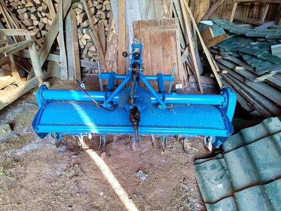 A blue rotavator/rototiller sitting in a barn along with some junk and firewood. It has a PTO input shaft and a three point hitch. Many curved tines lurk underneath its steel skirt.