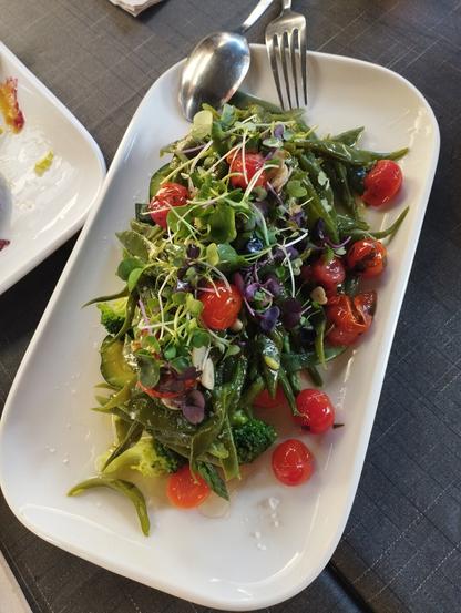Una ensalada vibrante y colorida servida en un plato blanco rectangular. Contiene una base de vegetales verdes como tiras largas y planas (posiblemente judías marinas o algas), floretes de brócoli y rodajas de calabacín. Se intercalan tomates cherry rojos, algunos ligeramente asados, y está coronada con una mezcla de microgreens verdes y morados, espolvoreada con sal marina y pequeños frutos secos (posiblemente piñones o almendras). Una cuchara y un tenedor de metal plateado descansan en la parte superior del plato. Se ve parte de una mesa oscura y un segundo plato blanco parcialmente comido en el fondo.