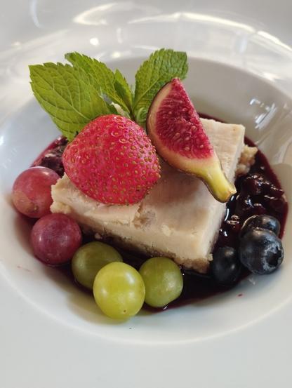 La imagen muestra un postre cuidadosamente emplatado en un plato blanco. El elemento central es una porción rectangular de un pastel o tarta de color crema claro, posiblemente un cheesecake, sobre una base más oscura. Este postre está rodeado por una vibrante salsa de frutos rojos oscura, que se extiende por la base del plato. Como guarnición, el plato incluye una variedad de frutas frescas y una hierba. En la parte superior, hay varias hojas de menta verde. A la derecha del postre, se encuentra una rodaja de higo con su interior rojizo y semillas visibles. Alrededor de la base del pastel, se observan una fresa entera de color rojo brillante, varios arándanos azules oscuros, y un grupo de uvas, tanto verdes como rojas.