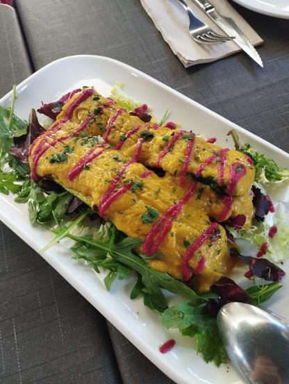 Una vista superior de un plato alargado y blanco con dos piezas principales de comida, servido sobre una mesa oscura. Las piezas son de forma oblonga, cubiertas con una salsa cremosa de color amarillo anaranjado brillante. Están decoradas con un hilo fino y vibrante de salsa rosa en patrón de zigzag y espolvoreadas con perejil fresco picado. Debajo de las piezas principales, hay una cama de ensalada fresca con hojas verdes variadas, como rúcula y algunas hojas de lechuga morada. En el fondo, se aprecian cubiertos plateados (un tenedor y un cuchillo) sobre una servilleta de papel beige, y parte de una cuchara plateada en la esquina inferior derecha del plato.