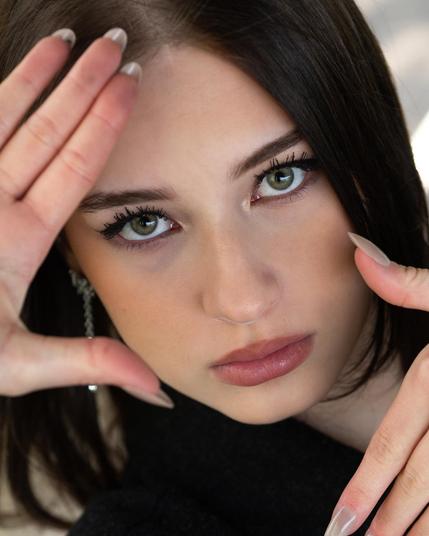 Woman with long dark hair and green eyes framing her face with her hands. Her nails are long and polished, and she wears earrings. The background is blurred, emphasizing her face.