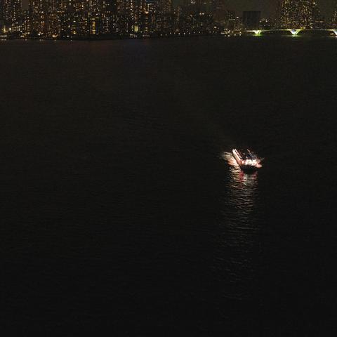 Boat illuminated by bright lights travels across a dark body of water, creating a bright path on the surface. City skyline with numerous lit windows is visible in the background. An arched bridge with glowing green lights spans part of the river.