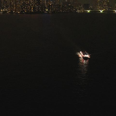 Boat illuminated by bright lights travels across a dark body of water, creating a bright path on the surface. City skyline with numerous lit windows is visible in the background. An arched bridge with glowing green lights spans part of the river.