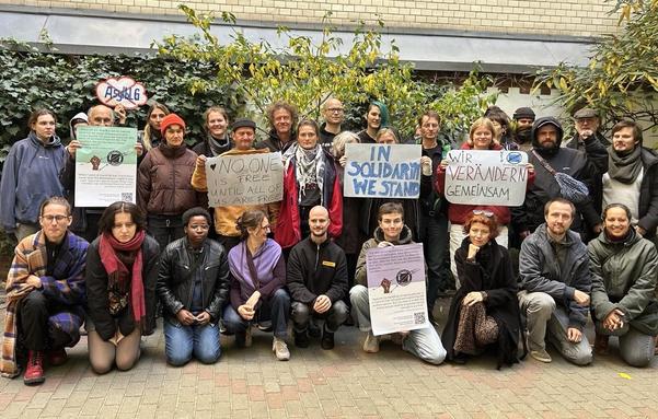 Gruppenbild von Teilnehmer*innen der Konferenz. Einige knien oder hocken in der vorderen Reihe, viele stehen dahinter.
Einige halten Schilder hoch. Auf den Schildern steht:
"No one is free until all of us are free", "In Solidarity we stand", "Wir verändern gemeinsam" und auf zwei Schildern ist nicht lesbarer Text mit einer Faust zu erkennen. Ein Schild hat die Form einer Wolke, auf der das Wort "AsylBLG" durchgestrichen ist.