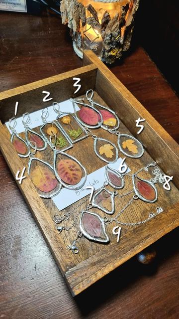 English A wooden drawer with stained glass earrings and bracelets containing autumn leaves and mushrooms. There are numbers next to each piece to choose from.
Français Un tiroir en bois contenant des boucles d'oreilles et des bracelets en vitrail contenant des feuilles d'automne et des champignons. Chaque pièce a un numéro à côté permettant de la choisir.