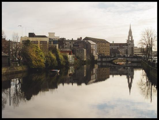 A serene view of the River Lee in Cork city in 2004 showing perfect mirror-like reflections of Georgian and Victorian buildings, a church spire, bridge, bare trees, and a small red boat on the completely still water under an overcast sky.