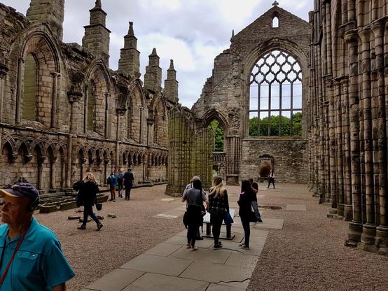 The ruins of Holyrood Abbey in Edinburgh, Scotland, feature weathered stone walls, tall arched windows, and partially collapsed columns, creating an open-air interior where the roof once stood. Visitors walk along the gravel and stone paths, some reading informational plaques while others explore the historic architecture. The left wall displays a series of repeating Romanesque arches and windows, while the far end opens to a large Gothic traceried window framing the greenery of the surrounding landscape. The space has a mix of tourists, some taking photos and others listening to audio guides, emphasizing its role as a popular historic site within the city.