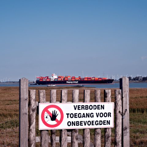 Photo of a wooden fence with a large "No Entry" sign, behind the sign the port of Antwerp is visible with a large container ship sailing by, the ship is centered in the image and framed by the poles of the fence.