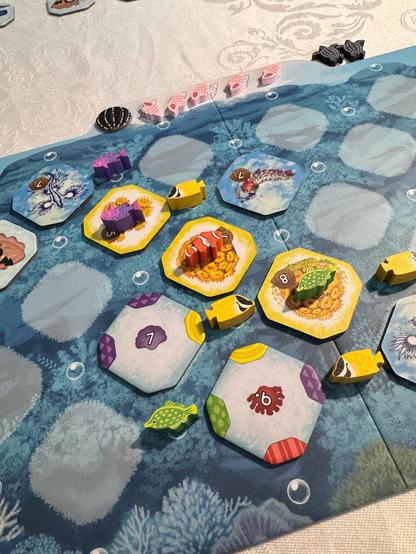 Das Bild zeigt einen Ausschnitt von einem Spielbrett. Das Brett hat einen blauen Hintergrund, der an Wasser erinnert, mit helleren und dunkleren blauen Wirbeln. Auf dem Brett befinden sich mehrere sechseckige Spielsteine. Die meisten Steine haben einen gelben Rand und ein farbiges Zentrum, das Muster enthält. Einige Steine sind lila, einige grün und einige rot. Auf einigen Steinen befinden sich kleine Spielfiguren, darunter eine Schnecke und eine Schildkröte. Auf dem Brett befinden sich auch kleinere blaue Spielmarker in Kreismuster. Einige Steine haben Zahlen darauf, einschließlich "7" und "8".