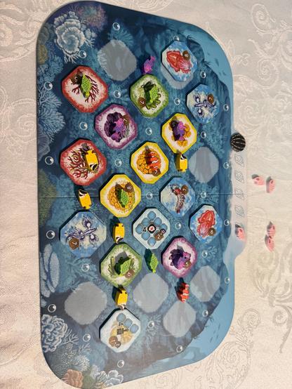 Eine schräge Luftaufnahme zeigt ein Spielbrett mit sechseckigen Spielsteinen. Das Brett ist länglich und mit einer dunkelblauen und grauen Struktur versehen, die an eine Landschaft oder eine Landkarte erinnert. Auf dem Brett sind 19 sechseckige Spielsteine in unterschiedlichen Farben und Mustern platziert. Die Spielsteine scheinen sich in einer unregelmäßigen Formation zu befinden, mit ein paar konzentrischen Ringen. Auf den Spielsteinen sind Zahlen von 4 bis 9 in kleinen schwarzen Ziffern zu sehen. Einige Spielsteine haben auch kleine Symbole, die einem Tier ähneln. Auf dem Brett befinden sich kleine, runde, weiße Markierungen, die über die Oberfläche verstreut sind.