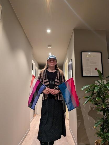 A gender-fluid person stands in a softly lit hallway holding two pride flags, one gender-fluid and one bisexual. They are wearing a green cap and a black dress. Framed art and a potted plant are visible in the background, adding a cozy and calm atmosphere.