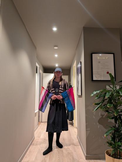 A gender-fluid person stands in a softly lit hallway holding two pride flags, one gender-fluid and one bisexual. They are wearing a green cap and a black dress. Framed art and a potted plant are visible in the background, adding a cozy and calm atmosphere.