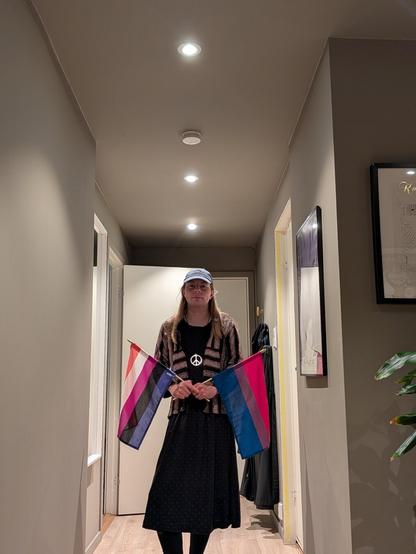 A gender-fluid person stands in a softly lit hallway holding two pride flags, one gender-fluid and one bisexual. They are wearing a green cap and a black dress. Framed art and a potted plant are visible in the background, adding a cozy and calm atmosphere.