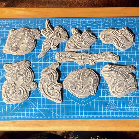 A blue cutting mat with nine small/medium sized Lino cuts laid out on it, they all appear to be medieval dragons (actually wyverns) or similar creatures inspired by manuscript maginalia