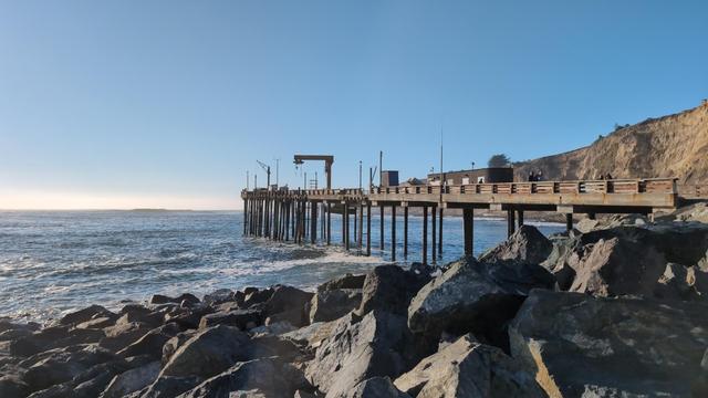 A wooden fishing pier in Point Arena Cove. Rocky shore in foreground, with the Pacific Ocean on the left side.