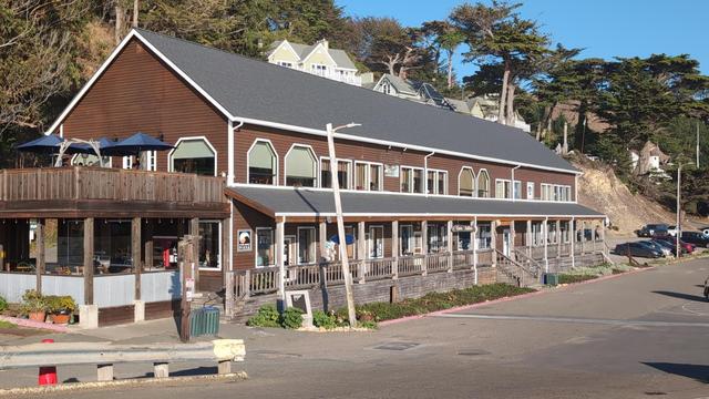 Wood building with full length porch. Parking lot can be seen on far right. Building contains many businesses, including a coffee shop, restaurant.