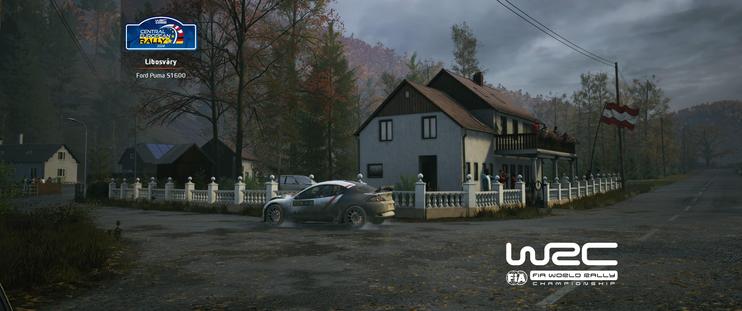 Alt text:

**A video game screenshot of a white and grey rally car, a Ford Puma S1600, sliding on a wet, paved area in front of a house in a rural setting.** The large, two-story house has a balcony and is surrounded by a white stone fence. A flag, possibly the flag of Austria, is visible on a pole near the house. The sky is overcast, and the surrounding trees show autumn foliage. In the upper left corner, there is a logo for the **"Central European Rally"** and the location **"Libešváry"** is listed. The lower right corner features the **"WRC FIA World Rally Championship"** logo.