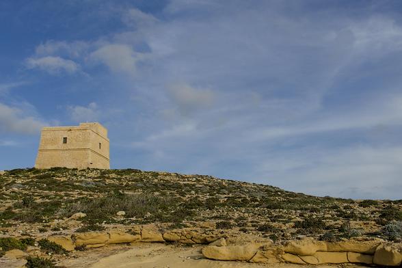 A small tower standing on a blue sky with sparse clouds. The ground has scrub grass and is a tan, rocky surface